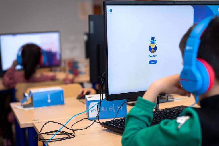A imagem mostra uma sala de aula de informática com crianças usando computadores. Um menino está sentado em frente a um monitor, usando fones de ouvido azuis. Na tela do computador, aparece o logotipo de um aplicativo chamado 'Portal'. Ao fundo, outra criança também está usando um computador. As mesas são de madeira e há caixas de som na mesa.