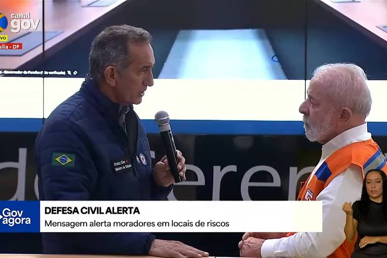 A imagem mostra uma reunião da Defesa Civil, onde duas pessoas estão conversando. Um homem, vestido com um colete laranja e cabelo grisalho, está ouvindo atentamente um outro homem que fala ao microfone, vestido com um casaco azul e com a bandeira do Brasil no peito. Ao fundo, há uma tela com informações e um logotipo do governo. Na parte inferior da imagem, há uma faixa com o texto 'DEFESA CIVIL ALERTA' e uma mensagem sobre alertas para moradores em locais de risco. Também há uma intérprete de libras à direita.