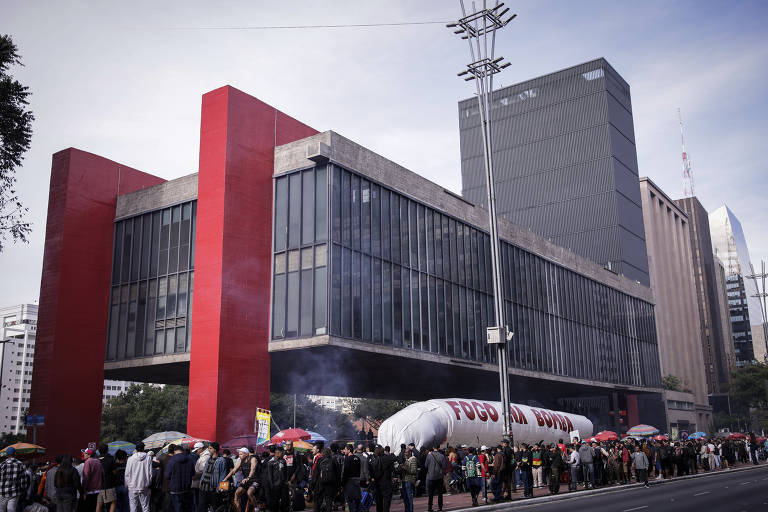 A imagem mostra uma grande multidão reunida em frente a um edifício moderno com uma fachada vermelha e janelas de vidro. As pessoas estão segurando guarda-chuvas coloridos e há uma faixa grande com a inscrição 'FOGO A BOLSA'. O ambiente parece ser de uma manifestação ou protesto, com fumaça visível no ar e outros edifícios altos ao fundo.