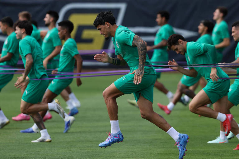 Um grupo de jogadores de futebol está em um campo, realizando exercícios de aquecimento. Eles estão vestidos com uniformes verdes e estão correndo em direção a um lado, puxando uma faixa elástica. O ambiente é ao ar livre, com grama verde e um fundo desfocado que sugere um campo de treinamento.