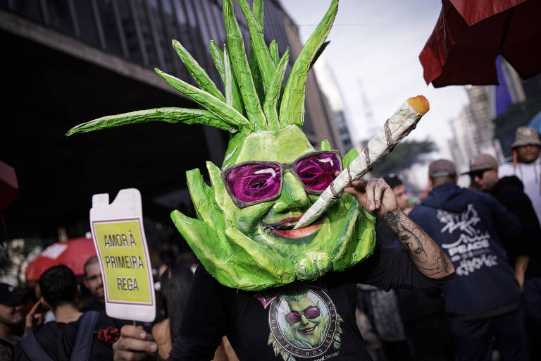 A imagem mostra um manifestante usando uma fantasia de cabeça de planta, com folhas verdes e óculos escuros. Ele segura um cartaz que diz 'AMOR À PRIMEIRA REGA' e tem um cigarro grande na boca. O fundo apresenta uma multidão e prédios, sugerindo um evento ao ar livre.