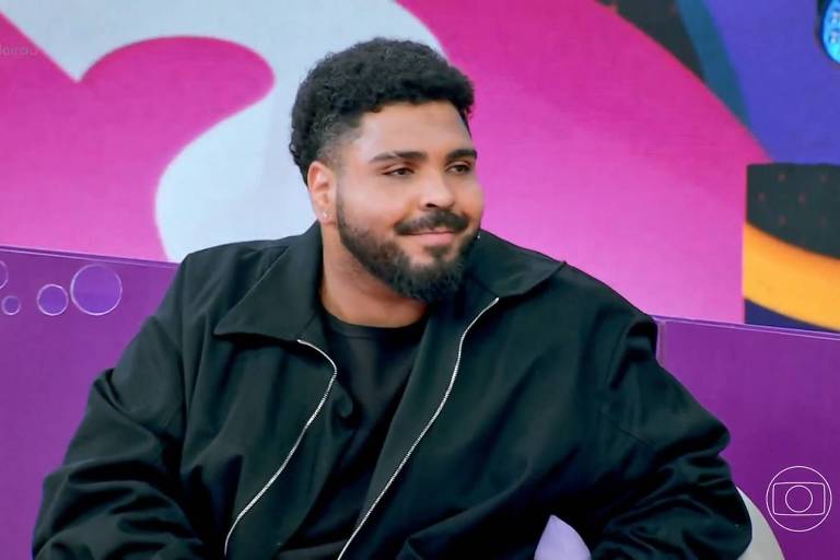 Um homem com cabelo crespo e barba está sentado em um sofá roxo. Ele usa uma jaqueta preta e uma camiseta preta. O fundo é colorido, com formas abstratas em tons de rosa e azul. O homem parece estar sorrindo levemente, em um ambiente descontraído de um programa de televisão.