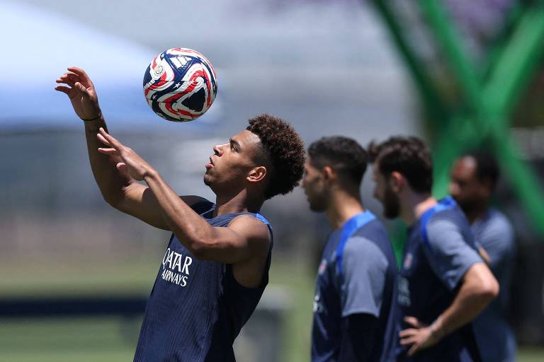 Um jogador de futebol está em um campo, levantando uma bola com as mãos. Ele usa uma camisa sem mangas e calças curtas, com chuteiras rosas. Ao fundo, outros jogadores estão parados, observando. O ambiente é ensolarado, com grama verde e uma estrutura de treinamento visível.