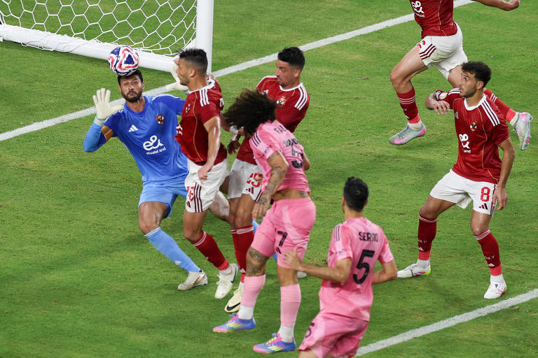 A imagem mostra um momento de uma partida de futebol, onde um goleiro, vestido com um uniforme azul, está prestes a defender a bola. Ele está cercado por jogadores de ambas as equipes, alguns vestindo uniformes vermelhos e outros em rosa. O goleiro, que é Mohamed El Shenawy, parece concentrado, enquanto os jogadores ao seu redor estão em posições variadas, indicando uma jogada intensa. O fundo mostra a rede do gol e o gramado do campo.