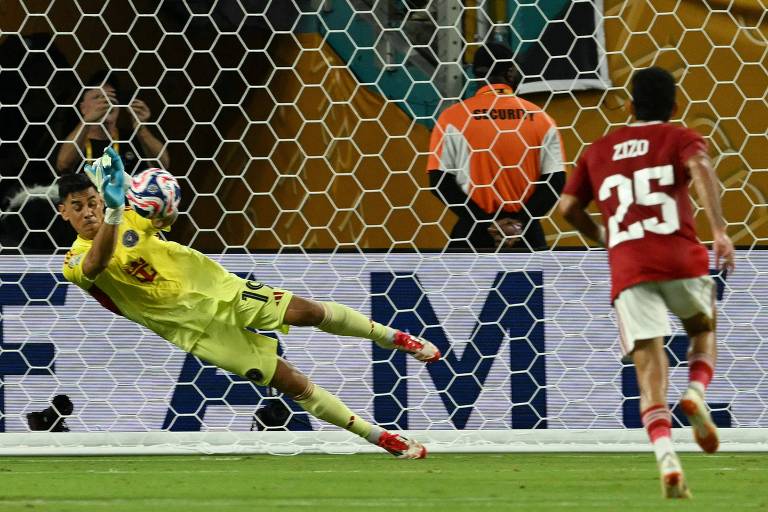 Um goleiro, vestido com um uniforme amarelo, está se esticando para defender um pênalti. Oscar Ustari está no ar, com a mão estendida para a bola, que está se aproximando. No fundo, há uma rede de gol e um jogador adversário, vestido com uma camisa vermelha e o número 25, observando a cobrança. O ambiente parece ser um estádio de futebol.