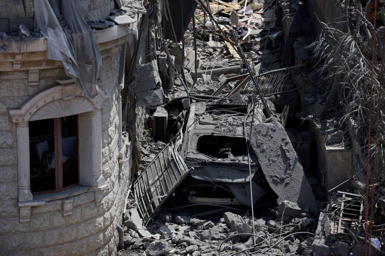 A imagem mostra uma área urbana devastada, com escombros e destroços visíveis. Há uma estrutura de pedra parcialmente intacta à esquerda, com uma janela visível. No centro, um veículo está coberto por detritos, e o chão está repleto de pedras e materiais de construção. Fios e estruturas metálicas estão expostos, indicando um colapso significativo na área.