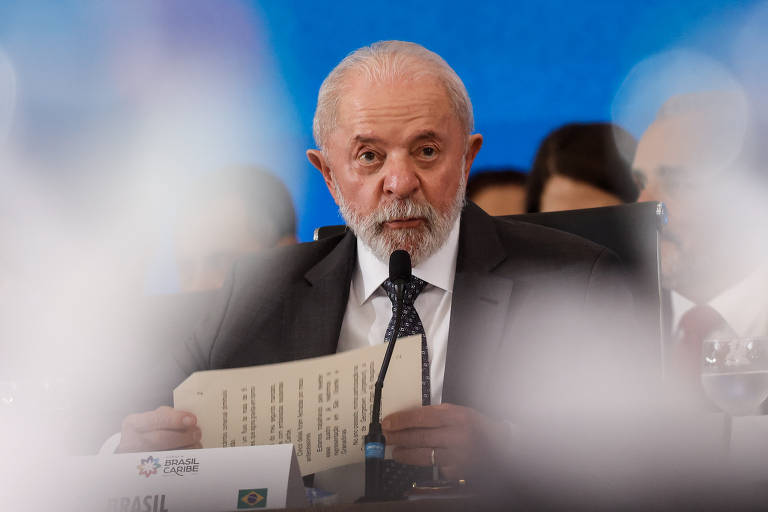 Um homem de cabelos grisalhos e barba, vestido com um terno escuro e gravata, está sentado em uma mesa durante uma conferência. Ele segura um documento em uma das mãos e fala ao microfone. Ao fundo, há uma tela azul e outras pessoas visíveis, mas desfocadas. Na mesa, há um copo de água e um pequeno suporte com a bandeira do Brasil.