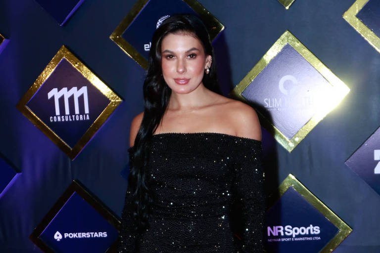 Uma mulher está posando em um tapete vermelho, vestindo um vestido longo preto com brilho. O fundo apresenta uma parede decorada com losangos em várias cores e logotipos de patrocinadores. A mulher tem cabelo longo e liso, e está sorrindo para a câmera.