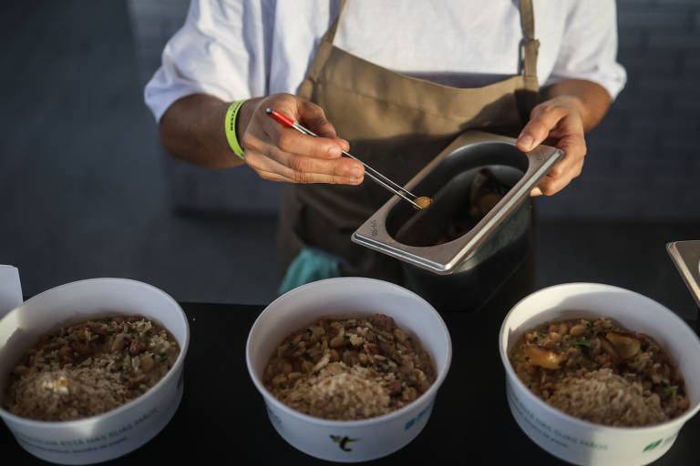 Um cozinheiro está servindo comida em três tigelas brancas. Ele usa um pincel para adicionar um molho ou tempero a uma das tigelas. O cozinheiro está vestindo uma camisa branca e um avental marrom, e há uma mesa com tigelas de arroz e outros ingredientes visíveis.