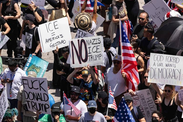 A imagem mostra uma multidão em um protesto, com várias pessoas segurando cartazes e bandeiras. Os cartazes contêm mensagens como 'ICE America's Gestapo', 'NO NAZIS ICE TRUMP', 'GOD FORBID A GIRL WANTS DEMOCRACY' e 'FUCK DONALD TRUMP'. A maioria das pessoas está vestida de preto e algumas usam bonés e óculos de sol. O ambiente parece ser urbano, com edifícios ao fundo.
