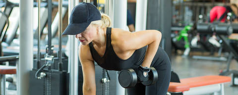 Beautiful mature woman in a black tracksuit suit and a cap does exercises with dumbbells in the gym. Sports training. (Foto: aleksandr_yu /Adobe Stock )