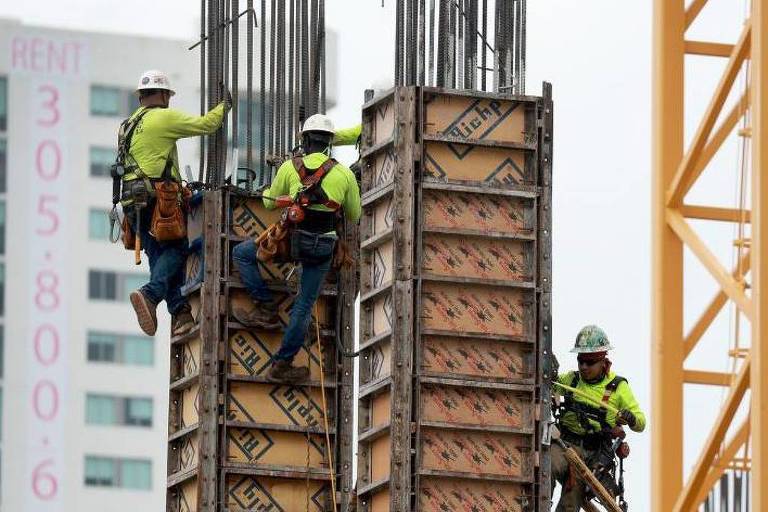A imagem mostra quatro trabalhadores da construção civil em um canteiro de obras. Eles estão usando coletes de segurança amarelos e capacetes brancos. Dois trabalhadores estão subindo uma estrutura de forma de concreto, enquanto os outros dois estão na parte inferior, próximos à base da estrutura. Ao fundo, é possível ver um edifício com uma placa de aluguel.