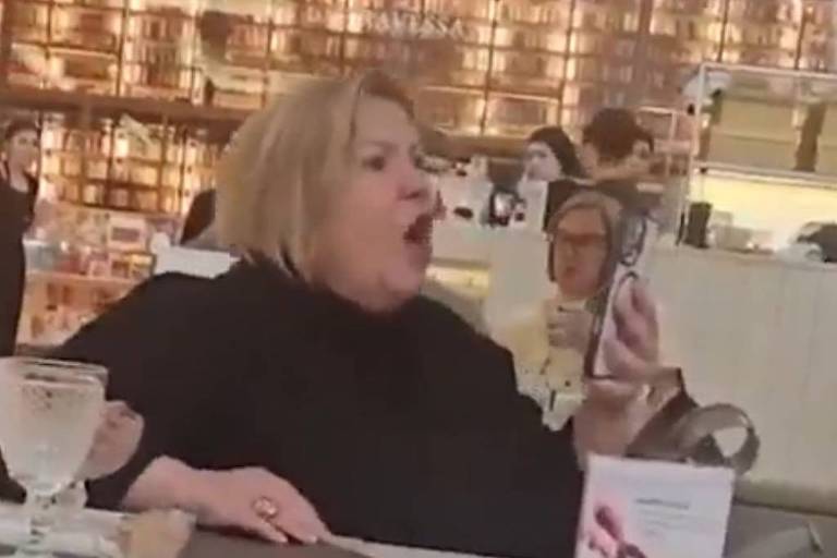 A imagem mostra uma mulher com cabelo loiro e curto, vestindo uma blusa preta, sentada em uma mesa de restaurante. Ela está gritando ou falando de forma expressiva, segurando um celular na mão. Ao fundo, há outras pessoas sentadas, algumas olhando para ela. A mesa tem copos e um cardápio visível.