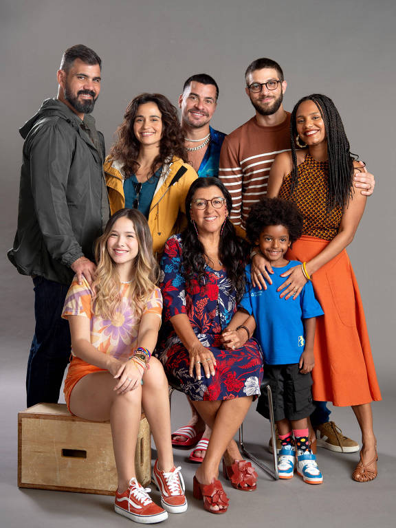 A imagem mostra um grupo de oito pessoas posando para a foto em um fundo cinza. Eles estão distribuídos em duas fileiras. Na frente, uma mulher de cabelo longo e liso, vestindo uma blusa colorida e shorts, está sentada em uma caixa de madeira. Ao lado dela, uma mulher mais velha, com cabelo curto e óculos, usa um vestido florido e está cercada por uma criança com cabelo cacheado e uma camiseta azul. Na parte de trás, há cinco homens e mulheres, alguns com cabelo curto e outros com cabelo longo, usando roupas casuais e coloridas. Todos estão sorrindo e parecem felizes.