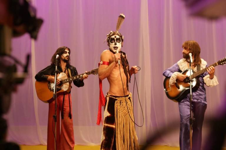 A imagem mostra três músicos se apresentando em um palco. O músico central está vestido com uma roupa exótica e pintado, segurando um microfone. Os outros dois músicos estão à sua esquerda e direita, tocando violões. O fundo é iluminado com luzes roxas e brancas, e há uma câmera visível em primeiro plano, sugerindo que a apresentação está sendo gravada.