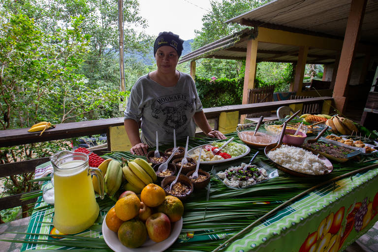 Uma mulher está em pé ao lado de uma mesa repleta de alimentos caseiros, incluindo frutas, saladas e pratos variados. A mesa está decorada com folhas verdes e há um jarro de bebida ao lado. Ao fundo, pode-se ver uma área verde com árvores e uma construção de madeira. A mulher usa uma camiseta e um lenço na cabeça, sorrindo para a câmera.