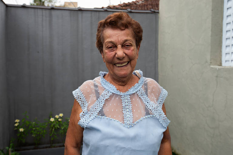 Uma mulher idosa está sorrindo para a câmera. Ela usa uma blusa azul clara com detalhes em renda. Ao fundo, há uma parede cinza e algumas plantas verdes. A mulher parece estar em um ambiente externo, possivelmente em um quintal ou jardim.