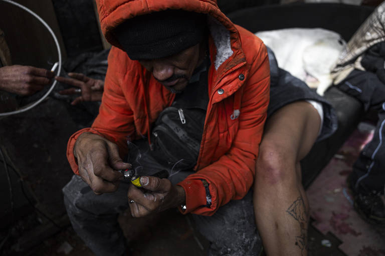 A imagem mostra um homem sentado em um sofá, vestindo um casaco laranja e uma touca preta. Ele está concentrado em seu cachimbo, enquanto uma mulher com uma tatuagem na perna está ao seu lado, parcialmente visível. O ambiente parece desordenado e escuro, com um cachorro branco deitado próximo a eles.