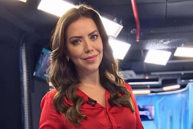 Uma mulher com cabelo longo e ondulado, vestindo uma blusa vermelha, está em um estúdio de televisão. O fundo é composto por luzes brilhantes e telas, sugerindo um ambiente de transmissão ao vivo. Ela está sorrindo e olhando para a câmera.