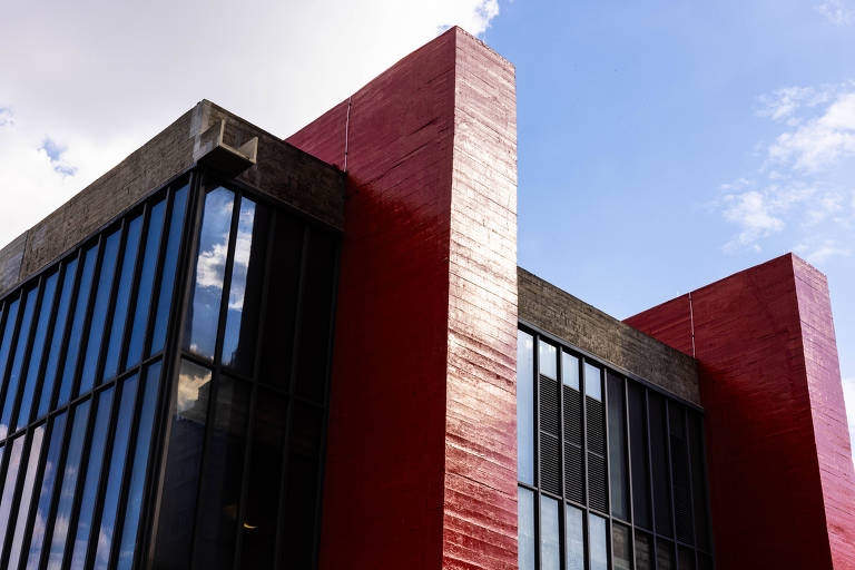 A imagem mostra a fachada de um edifício moderno, com paredes de concreto e grandes janelas de vidro. As paredes são predominantemente vermelhas, com uma textura brilhante, e há uma parte superior em concreto cinza. O céu está parcialmente nublado, com algumas nuvens visíveis.