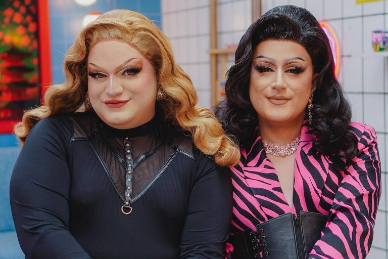 Duas drag queens estão posando juntas em um estúdio. A drag à esquerda tem cabelo loiro ondulado e usa uma blusa preta com detalhes em rede, além de calças jeans. A drag à direita tem cabelo preto liso e usa uma blusa rosa com listras pretas e um cinto largo. Ambas estão usando luvas pretas e estão sentadas em uma mesa, com um fundo de azulejos brancos e uma parede azul.