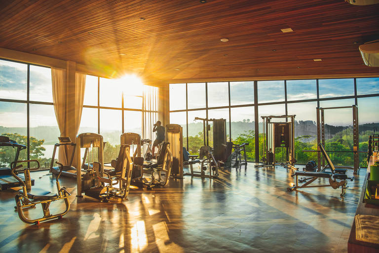 A imagem mostra o interior de uma academia com grandes janelas que permitem a entrada de luz natural. O sol está se pondo ou nascendo, criando um efeito de luz dourada que ilumina o espaço. Há vários equipamentos de ginástica, como máquinas de musculação e pesos, dispostos ao longo do ambiente. O piso é de madeira e há uma vista para a natureza ao fundo, com montanhas e vegetação visíveis através das janelas.
