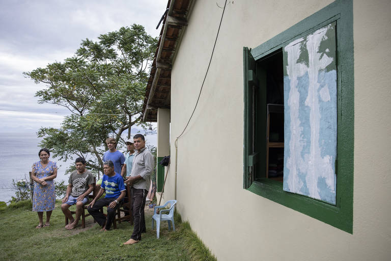 A imagem mostra um grupo de cinco pessoas em frente a uma casa de cor clara, com janelas verdes. Duas pessoas estão sentadas em cadeiras, enquanto três estão em pé. Ao fundo, há uma árvore e uma vista para o mar. O céu está nublado e a grama é verde.
