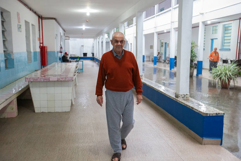 Um homem idoso caminha por um corredor amplo e iluminado, com paredes brancas e azuis. Ele usa um suéter vermelho e calças cinzas. Ao fundo, há outras pessoas e mesas dispostas ao longo do corredor. O piso é de cerâmica e há plantas decorativas em alguns pontos.