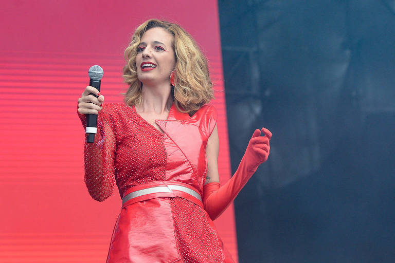 Uma mulher com cabelo loiro e ondulado está se apresentando em um palco. Ela usa um vestido vermelho brilhante com detalhes em strass e luvas vermelhas. A mulher segura um microfone com a mão esquerda e parece estar sorrindo enquanto canta. O fundo é de uma cor vermelha suave, criando um ambiente vibrante.