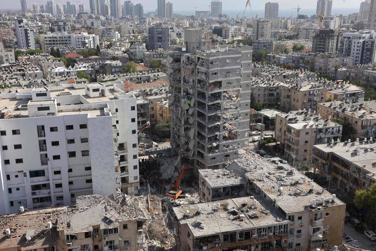 A imagem mostra uma área urbana devastada, com prédios danificados e em ruínas. Um edifício em destaque está parcialmente destruído, com escombros visíveis ao seu redor. Ao fundo, há uma vista da cidade com vários arranha-céus e construções. O céu está claro e a cena parece ser de um local que sofreu um desastre, possivelmente um ataque ou um terremoto.