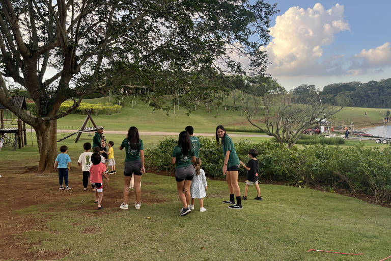 A imagem mostra um grupo de crianças e adolescentes em um parque. Algumas crianças estão brincando e explorando a área, enquanto adolescentes estão em pé, observando. Ao fundo, há uma grande árvore e uma paisagem verde com um céu parcialmente nublado. O ambiente parece ser tranquilo e ao ar livre.