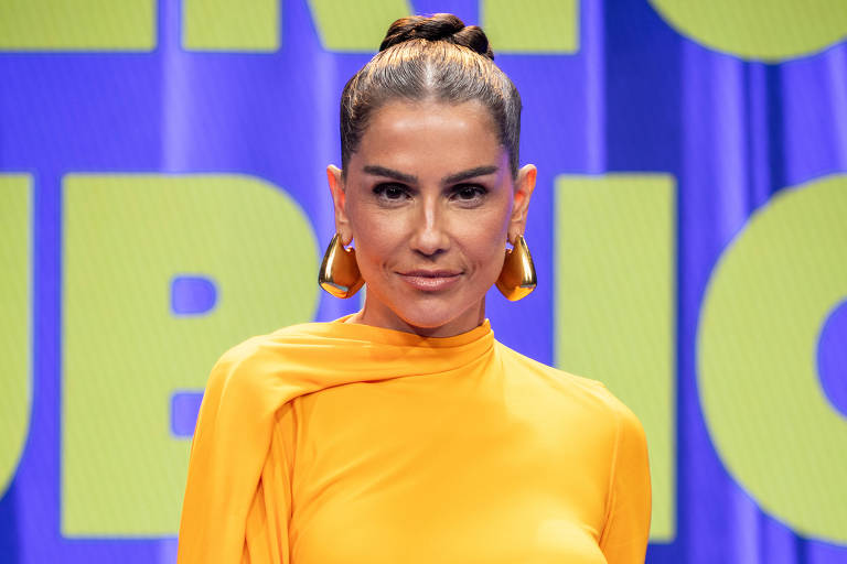 Uma mulher está em pé em um palco, vestindo um vestido longo e justo de cor laranja. O vestido tem um detalhe de manga longa que se estende até o chão. Ela está com o cabelo preso em um coque e usa brincos grandes. Ao fundo, há uma tela com a palavra 'PÚBLICO' em letras grandes e amarelas, e um fundo roxo.