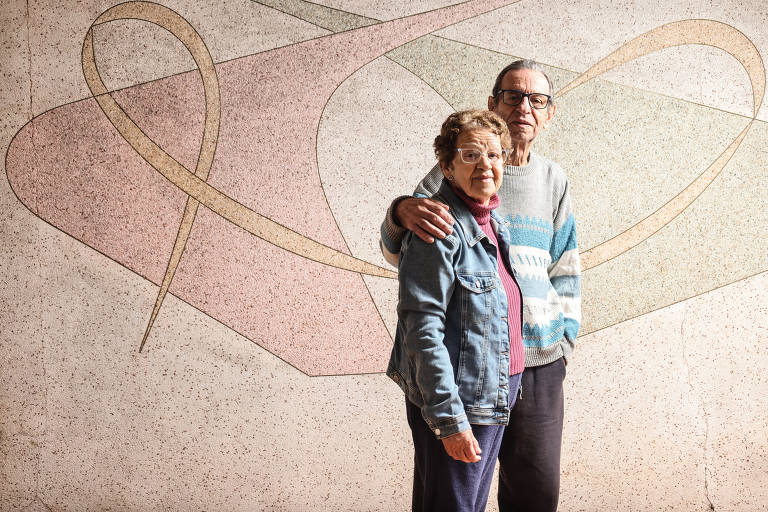 Um casal de idosos está posando para a foto em frente a um mural com formas geométricas em tons de rosa e bege. A mulher, com cabelo curto e óculos, usa uma jaqueta jeans e sorri, enquanto o homem, com cabelo grisalho e também usando óculos, está vestido com um suéter cinza e calças escuras. Ele coloca um braço ao redor dos ombros dela, demonstrando afeto.