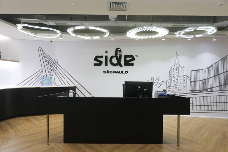 A imagem mostra a recepção de um espaço chamado Side em São Paulo. O balcão de recepção é preto e possui um computador em cima. Ao fundo, há uma parede branca com ilustrações em linhas de pontos turísticos de São Paulo, incluindo a Ponte Estaiada. O ambiente é moderno e bem iluminado, com luminárias circulares no teto.