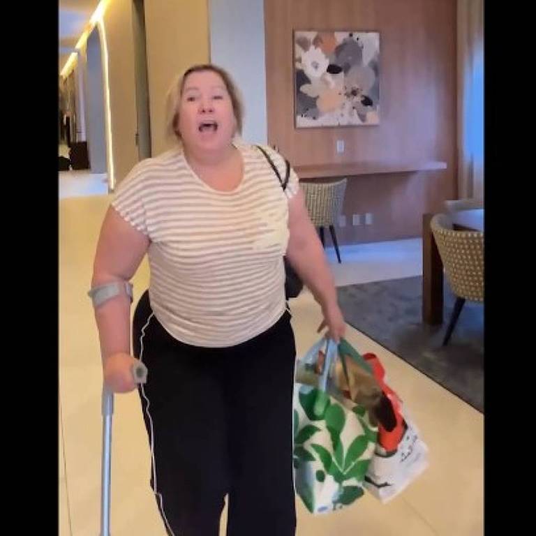 Uma mulher está em um ambiente interno, aparentemente em um hotel ou prédio comercial. Ela está usando uma camiseta listrada e calças escuras, segurando uma mala de rodinhas com uma mão e uma sacola com a outra. A mulher parece estar expressando uma emoção intensa, possivelmente raiva ou frustração. Ao fundo, há uma decoração simples com uma mesa e cadeiras, além de uma obra de arte na parede.