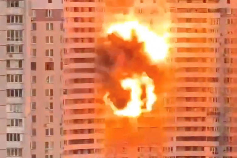 A imagem mostra um edifício residencial em chamas, com uma grande explosão visível em uma das janelas. A parte afetada do prédio está iluminada por uma intensa luz laranja e fumaça escura se espalha ao redor. O céu ao fundo é claro, indicando que é durante o dia.