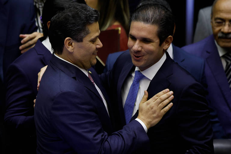 L'immagine mostra due uomini in abiti formali, apparentemente politici, che si stringono la mano e si sorridono. Uno dei due tiene la mano sul petto dell'altro, mentre il secondo sorride e sembra ricambiare il saluto. Sullo sfondo, sono visibili altre persone in abiti formali, ma non sono al centro dell'immagine.