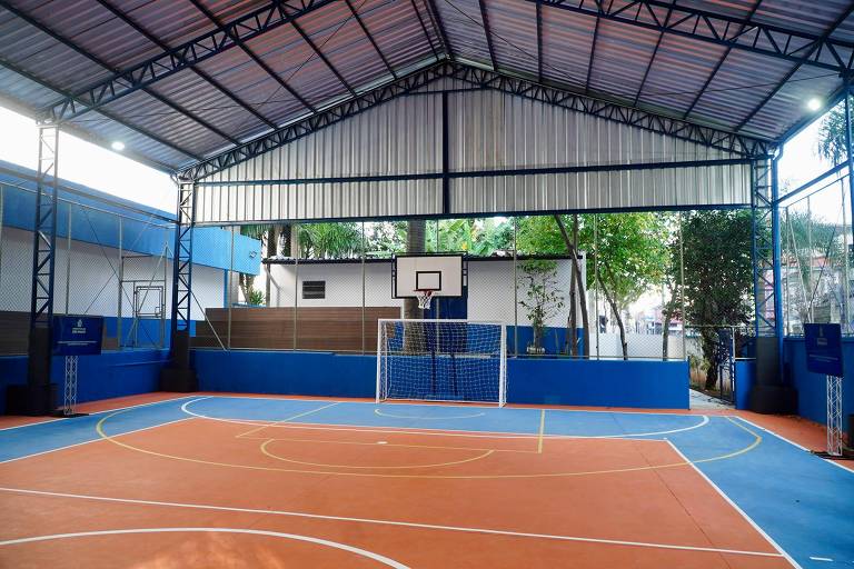 A imagem mostra uma quadra poliesportiva coberta com piso laranja e azul. Há uma cesta de basquete visível em uma das extremidades e um gol de futebol na outra. As paredes são de cor azul e há janelas que permitem a entrada de luz natural. Ao fundo, pode-se ver árvores e uma construção.