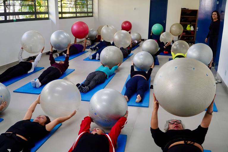 A imagem mostra um grupo de pessoas deitadas em colchonetes azuis, realizando exercícios com bolas grandes e prateadas. As pessoas estão em diferentes posições, segurando as bolas acima de suas cabeças. O ambiente é claro, com janelas que permitem a entrada de luz natural. Ao fundo, uma mulher observa a atividade.