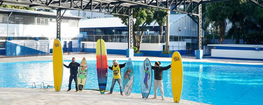 Cinco pessoas estão em pé ao lado de uma piscina, cada uma segurando uma prancha de surf. As pranchas variam em cores e designs, com algumas em tons de amarelo, azul e um gradiente colorido. O ambiente é coberto, com uma estrutura metálica visível acima e um fundo que mostra um espaço urbano. A piscina está vazia e reflete a luz do dia.