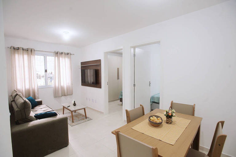 A imagem mostra um ambiente interno de um apartamento, com uma sala de estar e uma área de jantar. À esquerda, há um sofá cinza e uma mesa de jantar de madeira com cadeiras. A mesa está decorada com um caminho de mesa e uma fruteira. À direita, duas portas levam a quartos, e uma parede possui uma televisão montada. As janelas têm cortinas claras, permitindo a entrada de luz natural.