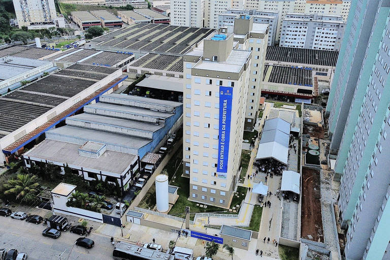 A imagem mostra uma vista aérea de um complexo urbano, com prédios residenciais e comerciais. No centro, há um edifício de vários andares com uma faixa azul na fachada. Ao redor, há várias construções de telhado metálico e uma área de estacionamento com veículos. O ambiente é urbano, com vegetação ao redor e outros prédios ao fundo.
