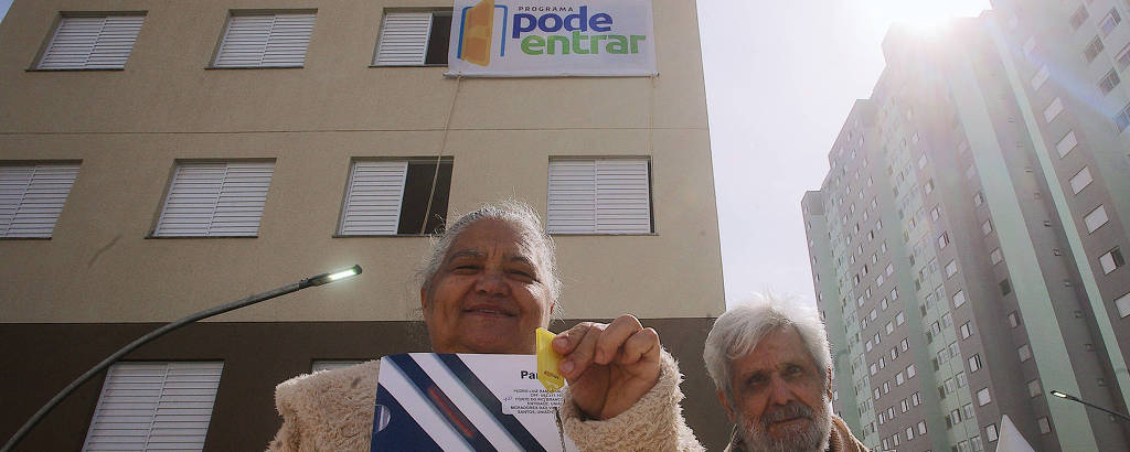 Uma mulher idosa sorri enquanto segura um documento e uma chave amarela, em frente a um prédio com uma faixa que diz 'pode entrar'. Ao lado dela, um homem idoso também está presente. O prédio é de cor clara e possui janelas. Ao fundo, há outros edifícios e uma tenda branca.