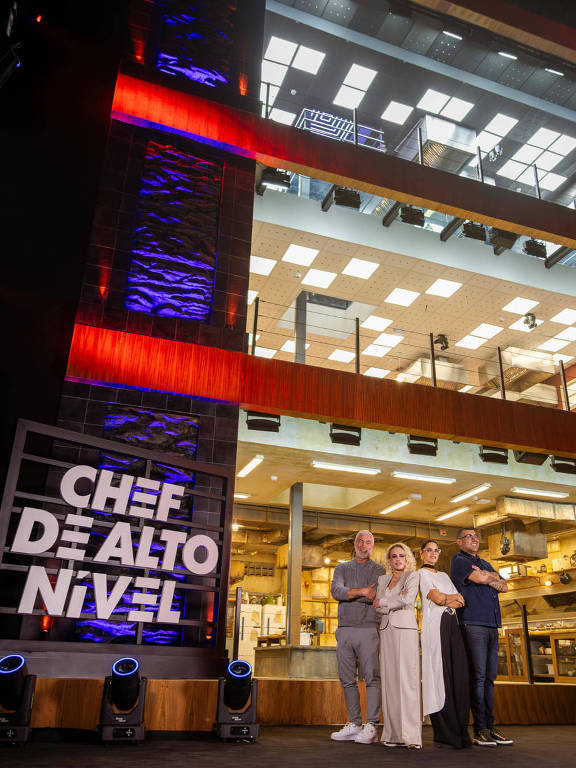 A imagem mostra quatro pessoas em frente a um grande letreiro que diz 'CHEF DE ALTO NÍVEL'. Ao fundo, há uma estrutura moderna com janelas amplas e iluminação interna visível. As pessoas estão posando para a foto, com expressões sorridentes, e vestem roupas casuais. O ambiente parece ser um estúdio ou um espaço de evento relacionado à gastronomia.