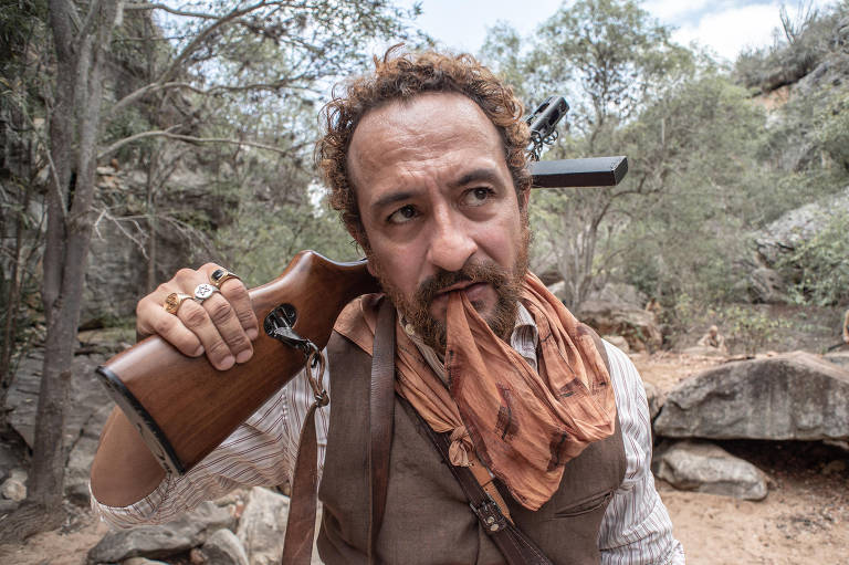 Um homem com cabelo encaracolado e barba, segurando um rifle em uma mão e um pedaço de tecido na boca. Ele está vestido com uma camisa clara e um colete, e usa anéis em uma das mãos. O fundo é uma paisagem natural com árvores e rochas.