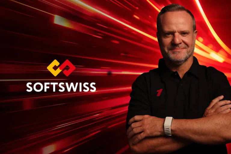 A imagem apresenta um homem sorridente com os braços cruzados, vestindo uma camisa preta com um logotipo vermelho e amarelo da SOFTSWISS ao lado. O fundo é uma explosão de luzes vermelhas e laranjas, criando um efeito dinâmico e energético. O homem usa um relógio branco no pulso esquerdo.