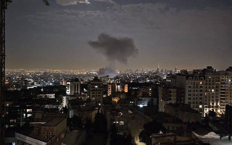 A imagem mostra uma vista noturna de uma cidade, com várias edificações visíveis. No céu, há uma nuvem de fumaça se elevando, indicando uma possível ocorrência de incêndio ou explosão. A cidade está iluminada por luzes, e ao fundo, é possível ver um horizonte com mais edifícios e uma leve iluminação. O céu apresenta algumas nuvens.