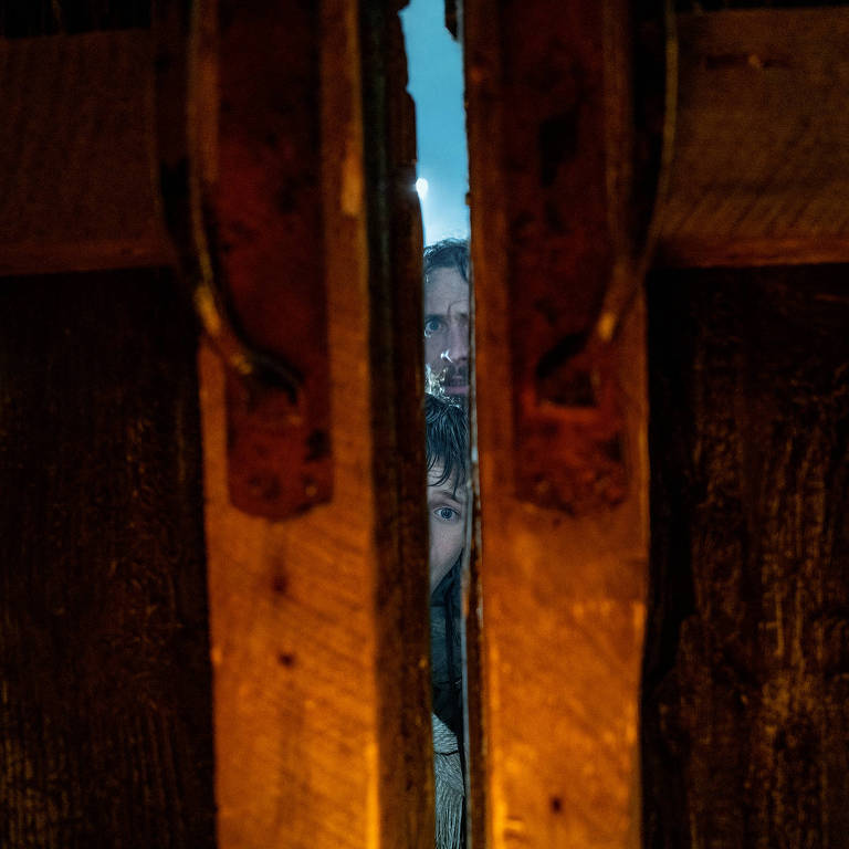 A imagem mostra uma visão através de uma porta de madeira, com tábuas verticais. Ao fundo, duas figuras humanas são visíveis, uma delas é uma criança e a outra parece ser um adulto. A iluminação é suave, criando um ambiente misterioso e intrigante.