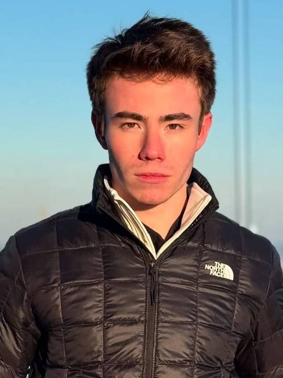 Um jovem está posando em uma plataforma elevada com vista para a cidade de Nova York. Ele usa uma jaqueta preta da marca The North Face e luvas. Ao fundo, é possível ver arranha-céus e uma paisagem urbana, com um céu claro e algumas nuvens.
