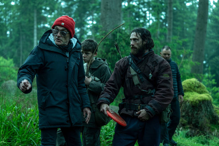 Um grupo de quatro pessoas está em uma floresta densa. Um homem mais velho, usando um gorro vermelho e um casaco escuro, está apontando com a mão. Ao seu lado, um jovem está segurando um arco. Outro homem, com uma expressão séria, segura um disco de plástico vermelho. Ao fundo, um quarto homem observa a cena. A vegetação ao redor é verde e exuberante, com árvores altas.